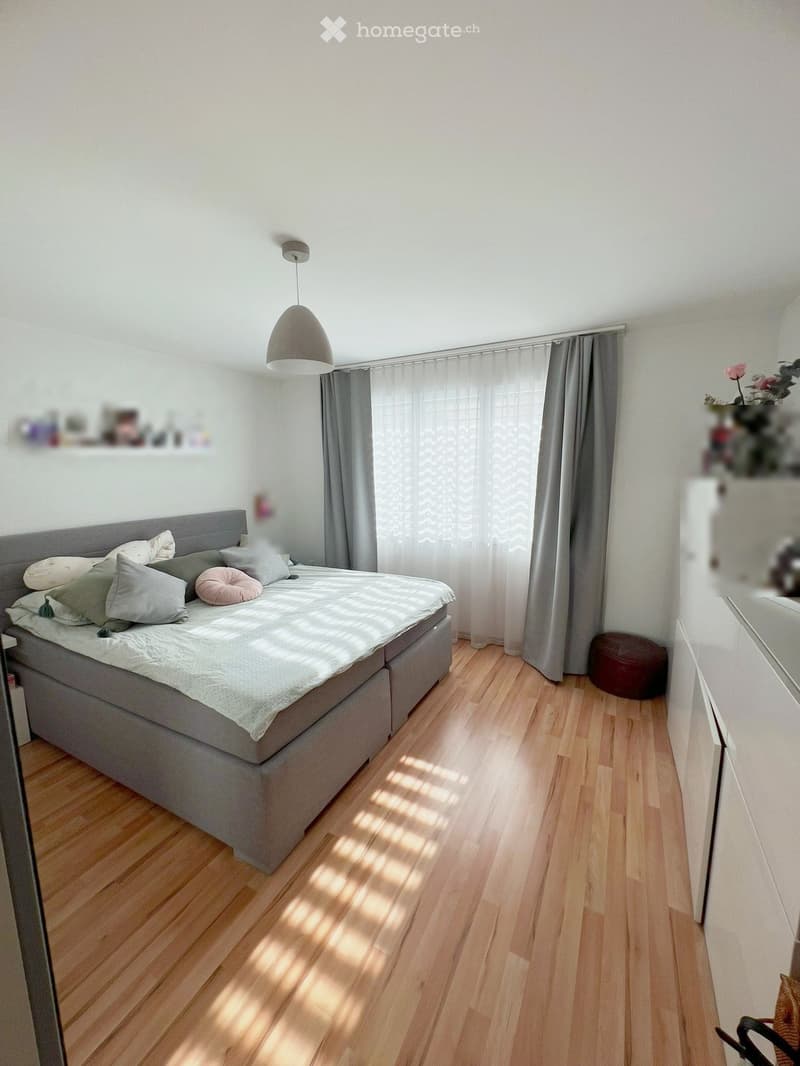 Bright bedroom with wooden flooring, a grey bed with multiple pillows, a hanging lamp, grey curtains, a small round stool, and a shelf with decorative items.