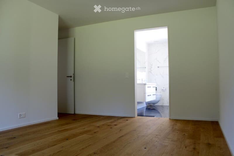 Empty room with light green walls and wooden flooring, featuring an open doorway leading to a modern bathroom with white fixtures.
