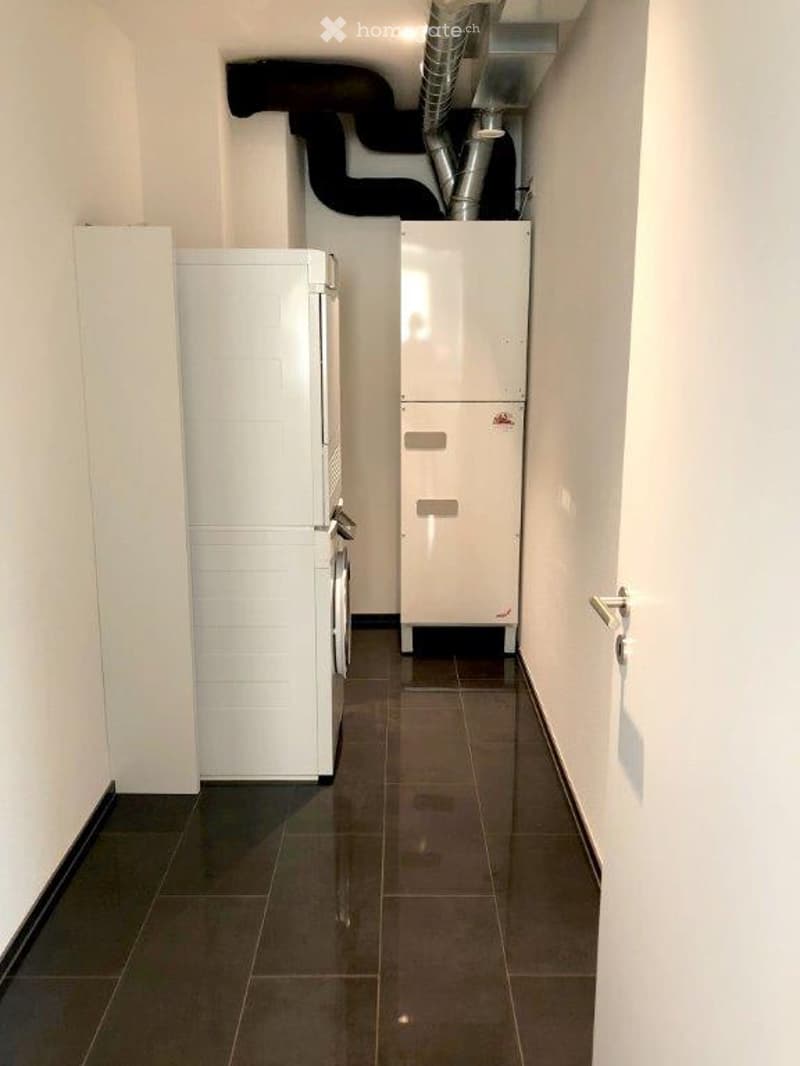 A narrow hallway with tiled flooring, white walls, and a white cabinet on the left side. There is a white appliance, possibly a washing machine, on the right side.
