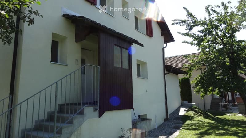 Two-story house with light-colored exterior, wooden entrance door, and small garden area with grass and gravel.
