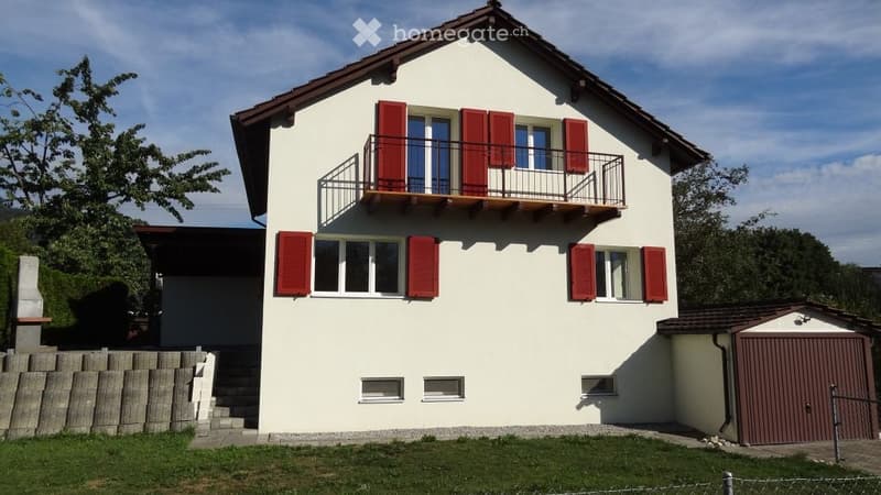 Two-story house with white walls, red shutters, and a balcony on the upper floor. Features a garage and a small garden area.