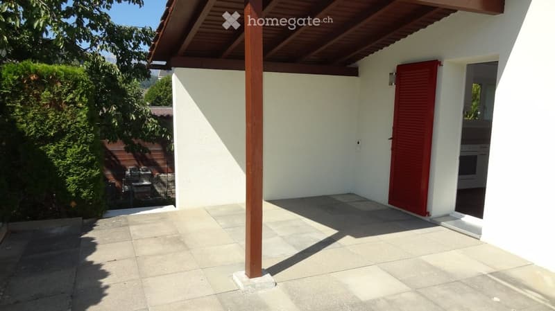 A covered patio area with a tiled floor, white walls, and a red shuttered door leading to the interior. There is a wooden support column and a view of a garden with a grill and outdoor furniture.