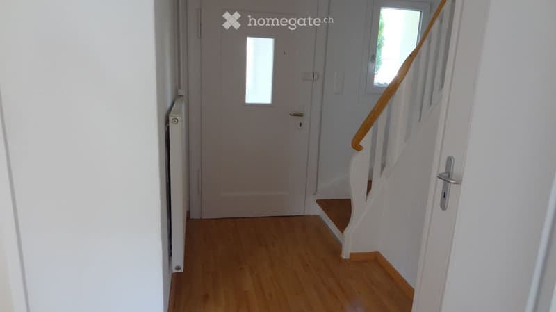 A narrow hallway with wooden flooring, white walls, a staircase with a wooden handrail, and a closed door with a small window.