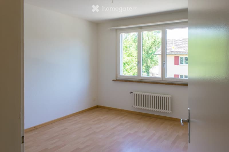 Empty room with light wooden flooring, white walls, a radiator, and a large window with a view of greenery and a neighboring building.