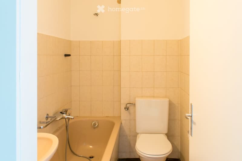 Small bathroom with a bathtub, toilet, and sink. Light-colored tiles and a simple design.