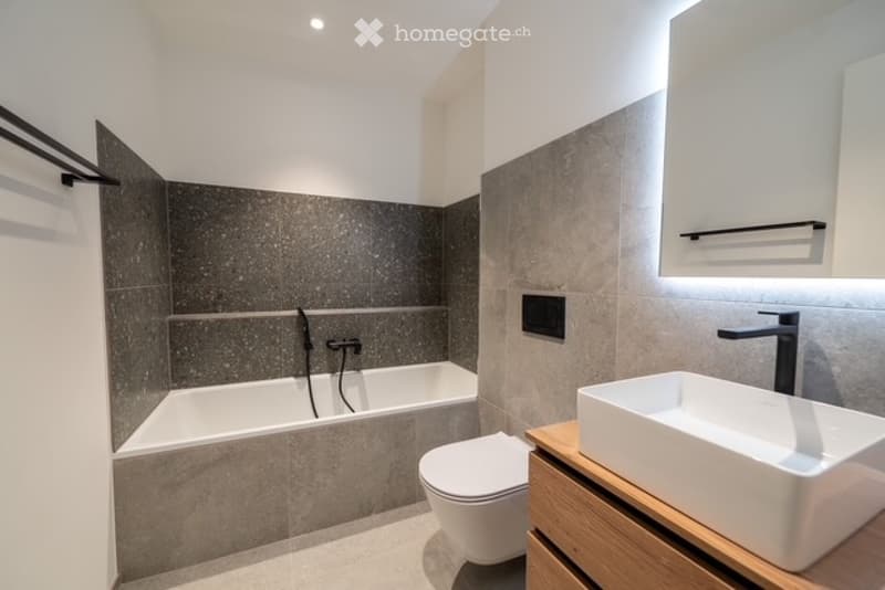 Modern bathroom with a bathtub, toilet, and sink. Features grey tiled walls, a wooden vanity, and sleek fixtures.
