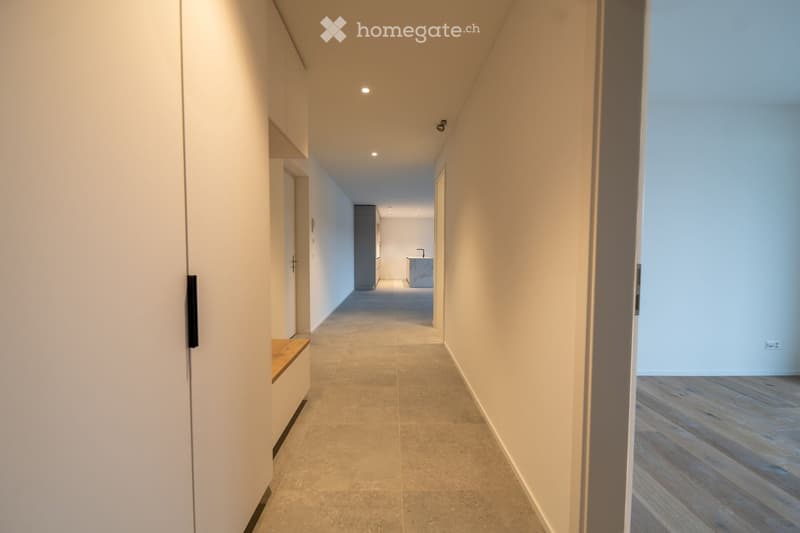 Modern hallway with light-colored tiled flooring, white walls, built-in storage units, and recessed lighting. The hallway leads to a room with a wooden floor and a visible kitchen area in the distance.