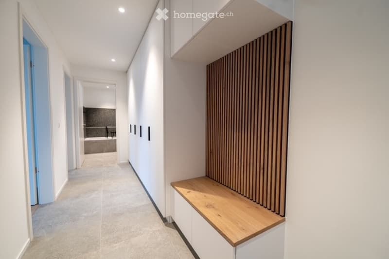 Modern, minimalist hallway with white walls, built-in wooden bench, and open shelving unit. The hallway leads to a bathroom with a visible bathtub and shower area.