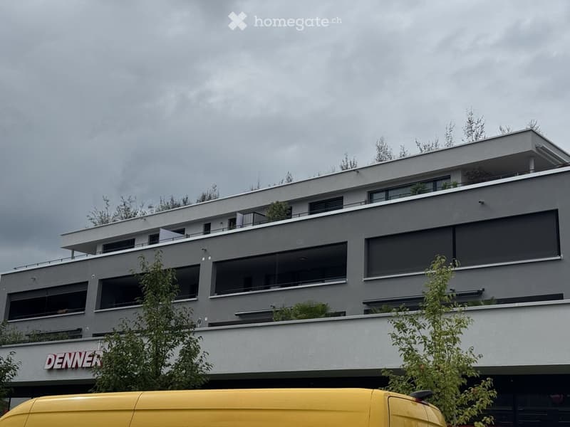 Modern multi-story building with large windows, balconies, and greenery on the roof and surrounding areas.