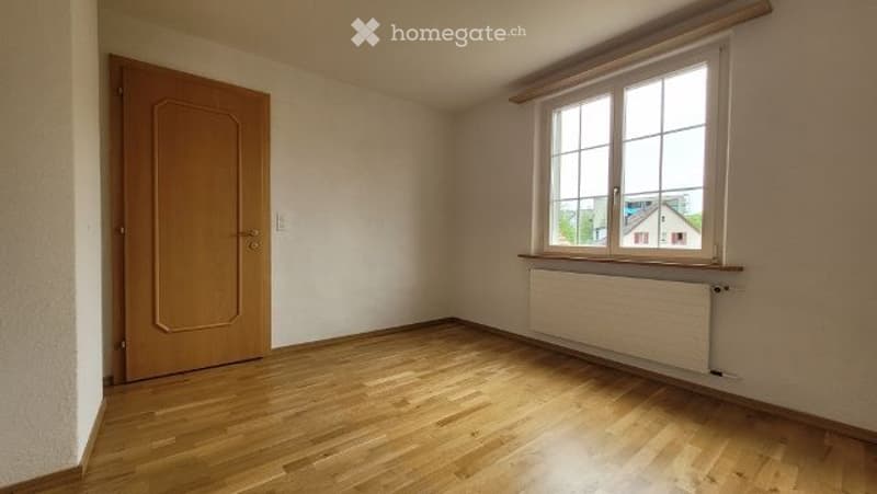 Empty room with wooden flooring, white walls, a window with blinds, and a radiator below it. The room has a wooden door on one side.