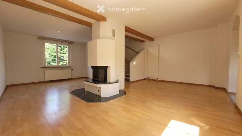 Bright, unfurnished room with wooden flooring, white walls, exposed wooden beams, and a central fireplace. Natural light streams in from a large window on one side.