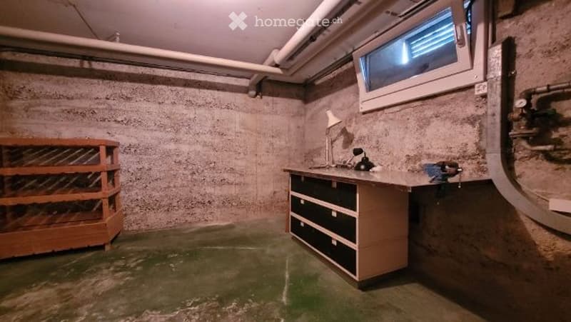 The room has a concrete wall finish, a wooden shelf on the left, a desk with drawers, and a small lamp on the desk. There is a window at the top right corner, and the floor appears to be concrete.
