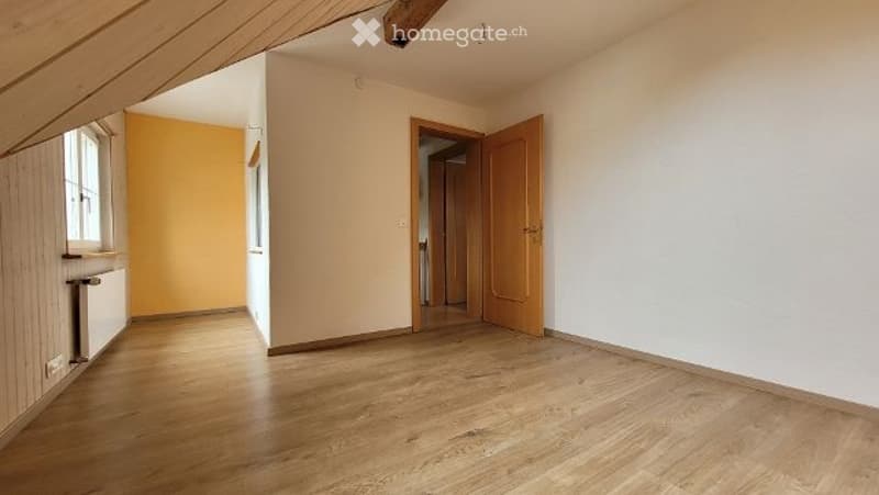 Empty room with wooden flooring, white walls, and a slanted ceiling typical of an attic space. Features include a window with wooden frame, a radiator, and an open doorway leading to another room.