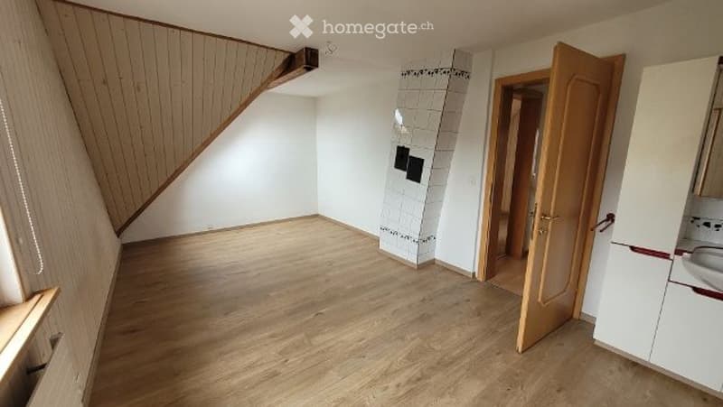 Empty room with wooden flooring, white walls, and a staircase leading upwards. A doorway leads to a tiled bathroom area with a sink visible.