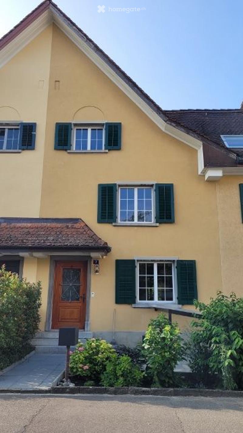 Two-story yellow house with green shutters, wooden front door, and small garden area.