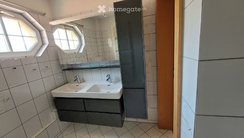 Modern bathroom with white tiled walls and floor, double sink vanity with dark cabinets, large mirror, and two octagonal windows providing natural light.