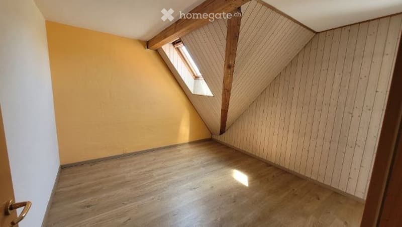 Empty attic room with wooden floor, slanted wooden ceiling beams, and a small skylight window letting in natural light.