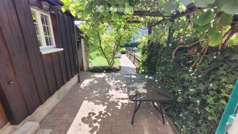 A narrow garden path leads to a small outdoor seating area with a table, surrounded by lush greenery and a wooden house on one side.