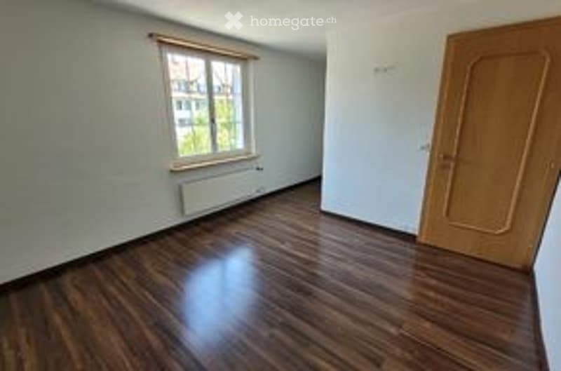 Empty room with wooden flooring, white walls, a window with a view, and a closed wooden door.
