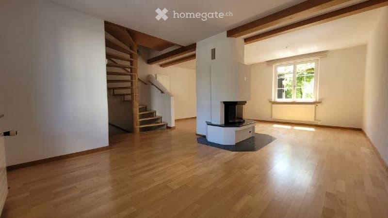 Empty room with wooden flooring, white walls, exposed wooden beams on the ceiling, a staircase leading to an upper level, and a fireplace in the corner.