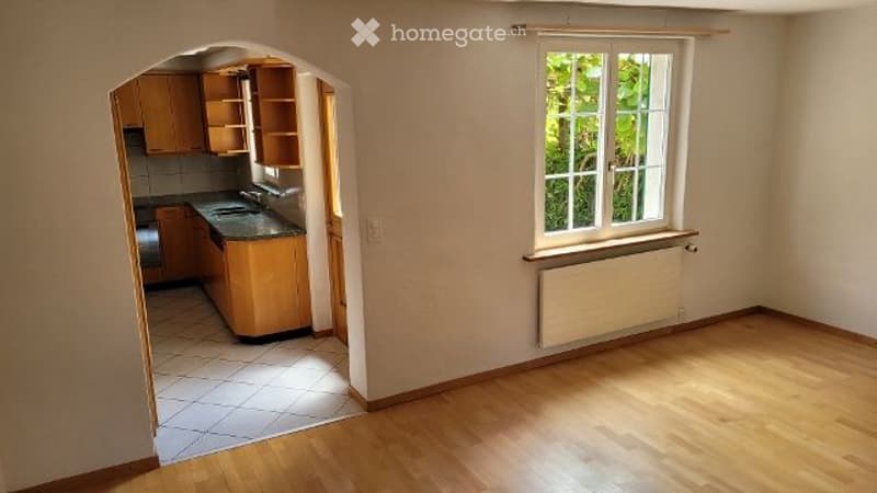 Empty living space with wooden flooring, white walls, and a doorway leading to a small kitchen area with cabinets and a sink.