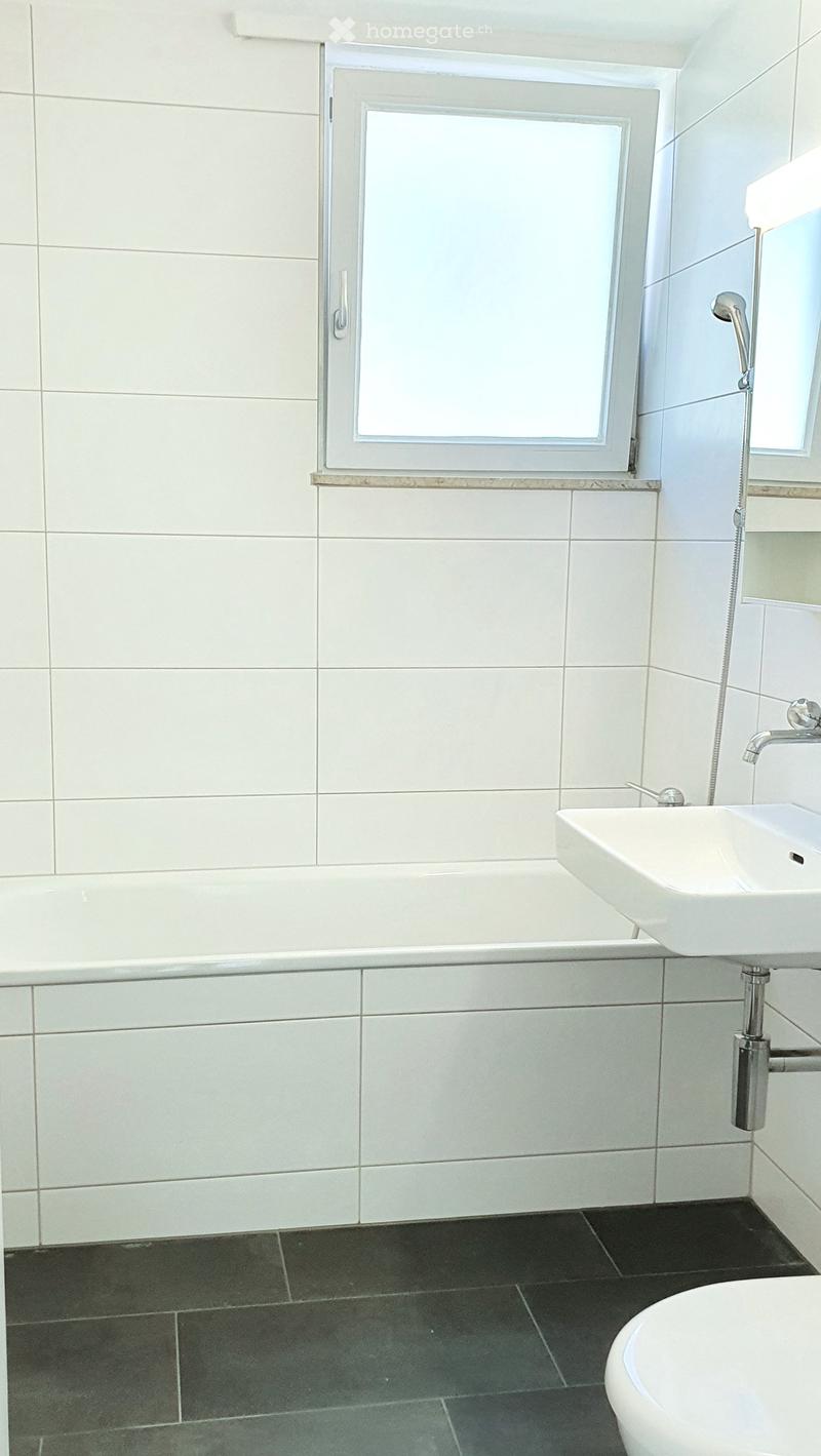 Modern bathroom with white tiled walls, a bathtub, a sink with a chrome faucet, and a toilet. The floor is dark grey tiles, and there is a window above the bathtub.