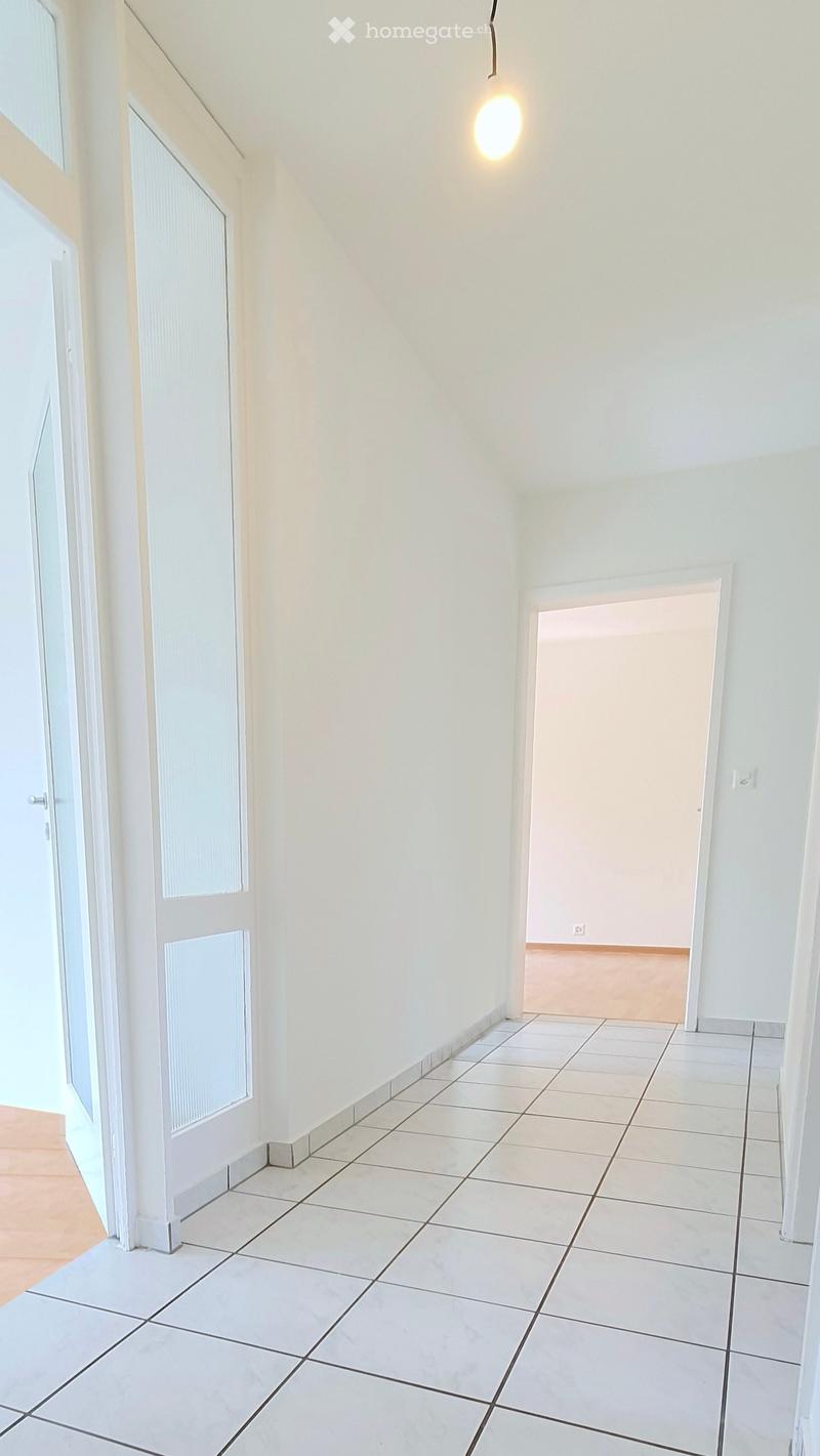 Empty hallway with white tiled flooring, white walls, and a single hanging light fixture. Two doors are visible, one leading to an open area and the other to a closed room.