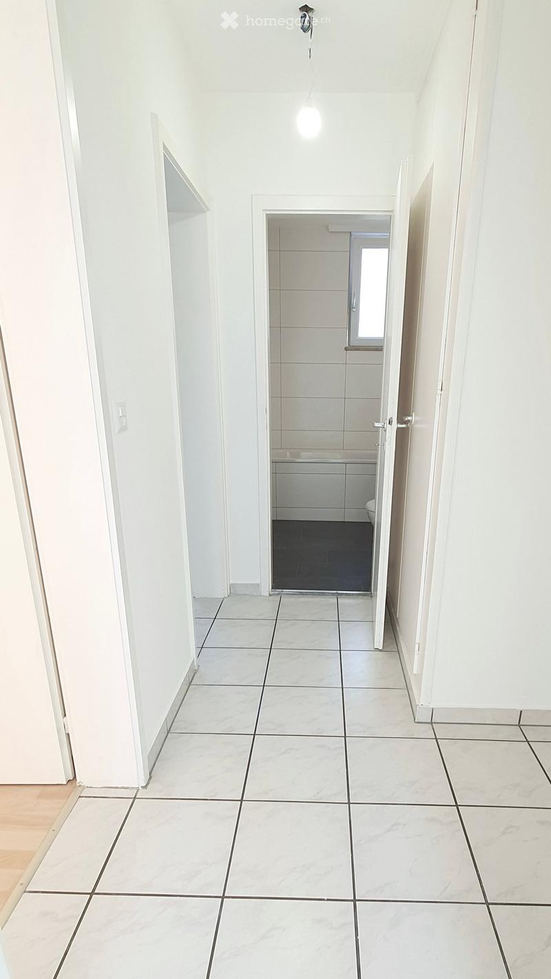 A clean, white-walled hallway with marble tiles, leading to a small bathroom with a toilet and sink.