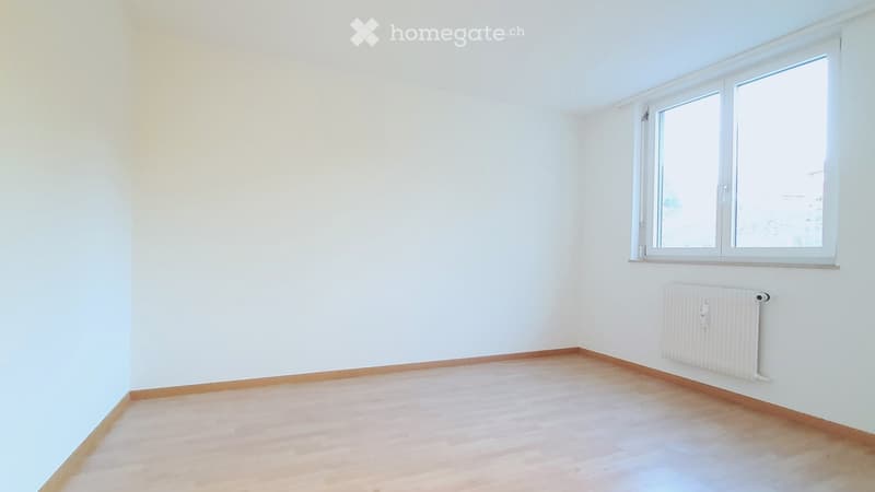 Empty room with white walls, wooden flooring, and a window with white frame. A radiator is mounted on the wall below the window.