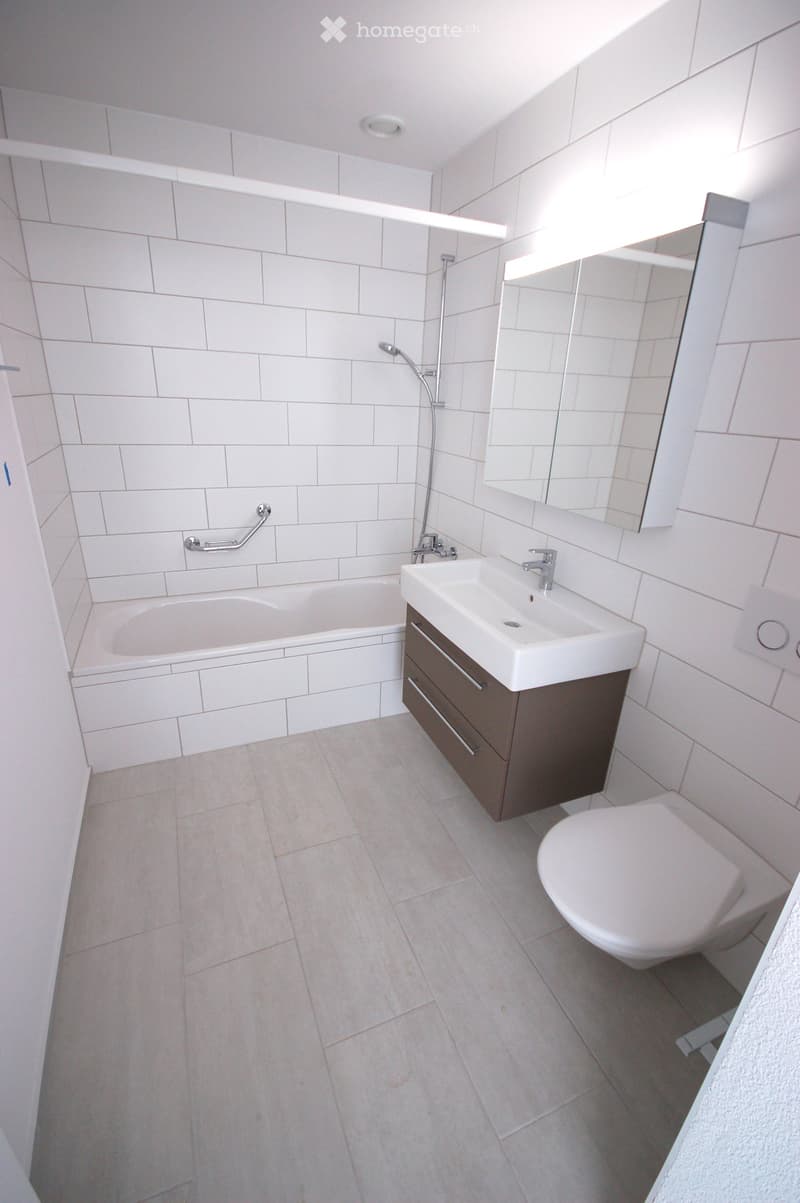 Modern bathroom with white tiles, bathtub, sink, toilet, and a mirrored cabinet.