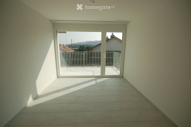 Empty room with tiled floor, white walls, and large sliding glass doors leading to a balcony with a metal railing and a view of rooftops and distant hills.