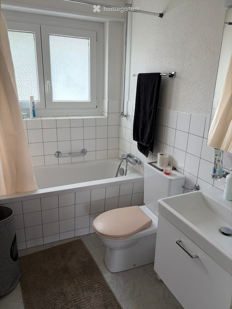 Modern bathroom with white tiles, bathtub, toilet, sink, towel, and toiletries.