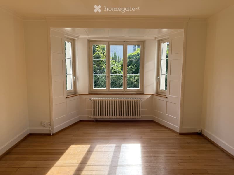 Empty room with light-colored walls, wooden flooring, and large windows allowing natural light to enter. A radiator is visible below the central window.