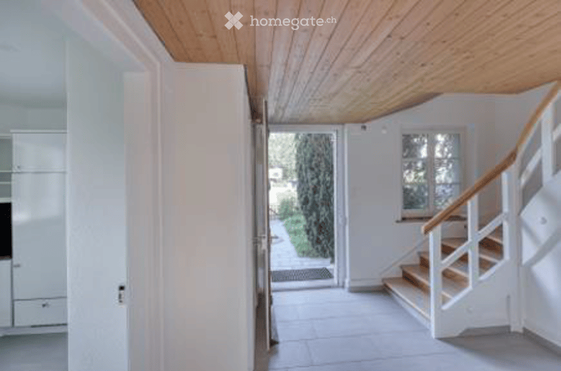 Bright hallway with white walls, wooden ceiling, tiled floor, and a staircase leading to an upper level. A door opens to a garden area with greenery visible.