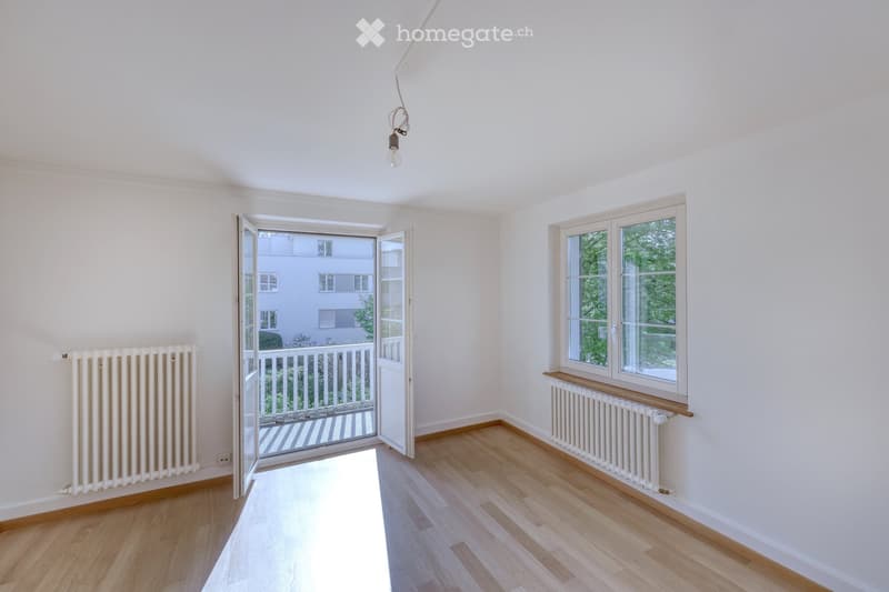 Empty room with light wooden flooring, white walls, and large windows. Features a radiator on each wall and an open sliding door leading to a balcony.