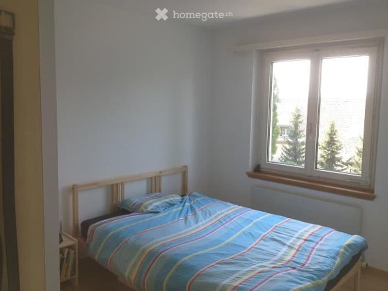A small bedroom with a single bed, blue and green striped duvet, a pillow, and a nightstand next to the bed. The room has light blue walls and a window with white frames.