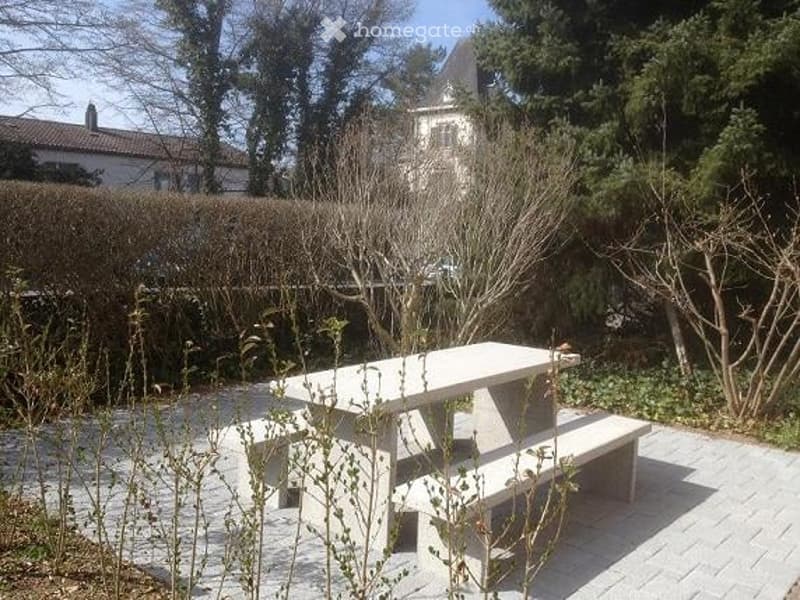 A small outdoor seating area with a concrete picnic table and benches, surrounded by low hedges and trees, with a house visible in the background.