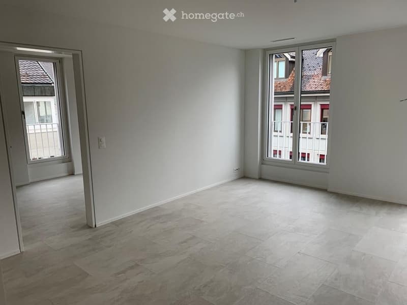 Empty room with light-colored walls, tiled floor, and large windows offering views of neighboring buildings.