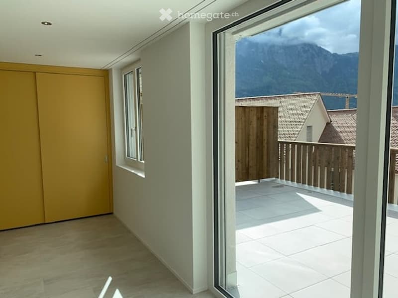 Bright, unfurnished room with wooden flooring, large windows, and an open sliding door leading to a tiled balcony with a scenic mountain view.