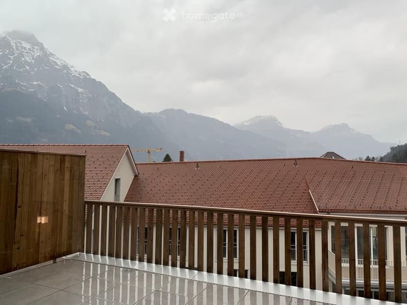 Balcony with wooden railing overlooking a building complex and distant mountain range.