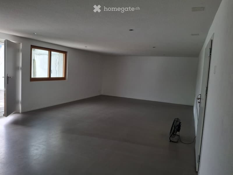 Empty room with bare walls, concrete floor, and a single window with wooden frame. A small fan is placed in the corner.