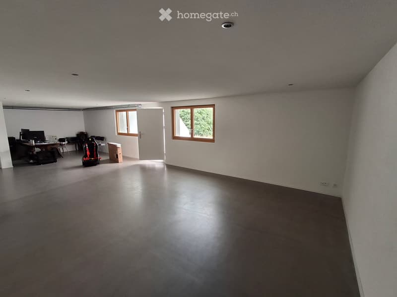 A large, open room with a polished concrete floor, white walls, and two windows. The room contains a desk with a computer and a gaming chair, along with a few other chairs and a small table.