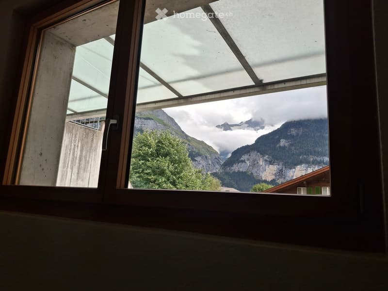 View from a balcony or terrace showing a scenic mountain landscape with trees and a house in the foreground.