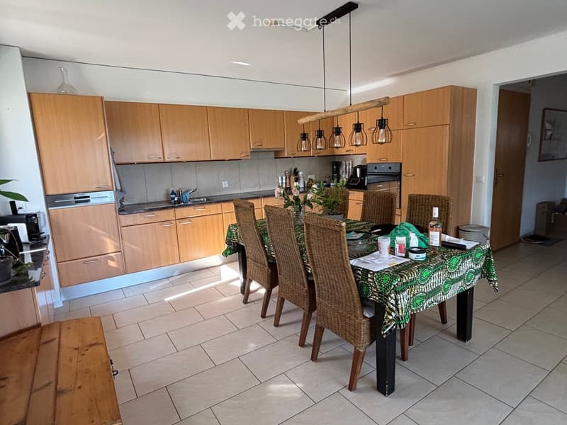 Bright kitchen with light wooden cabinets, a central dining table with wicker chairs, and various appliances including a sink, oven, and dishwasher.