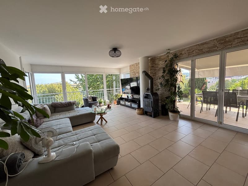 Bright living room with large windows and sliding glass doors leading to a balcony. Features a grey sectional sofa, coffee table, fan, TV, and additional seating.