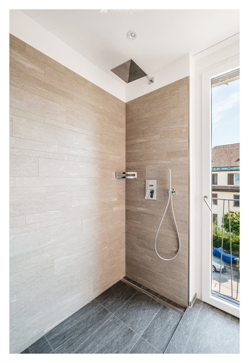 Modern shower area with light wood tile walls, dark grey tiled floor, and a glass door leading to a balcony.