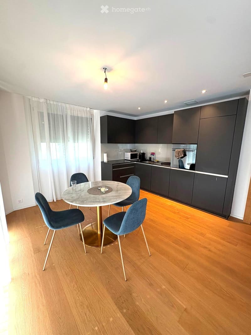 A modern, well-lit room with a round marble table and four blue chairs, a kitchen area with dark cabinets and a microwave, and large windows with white curtains.