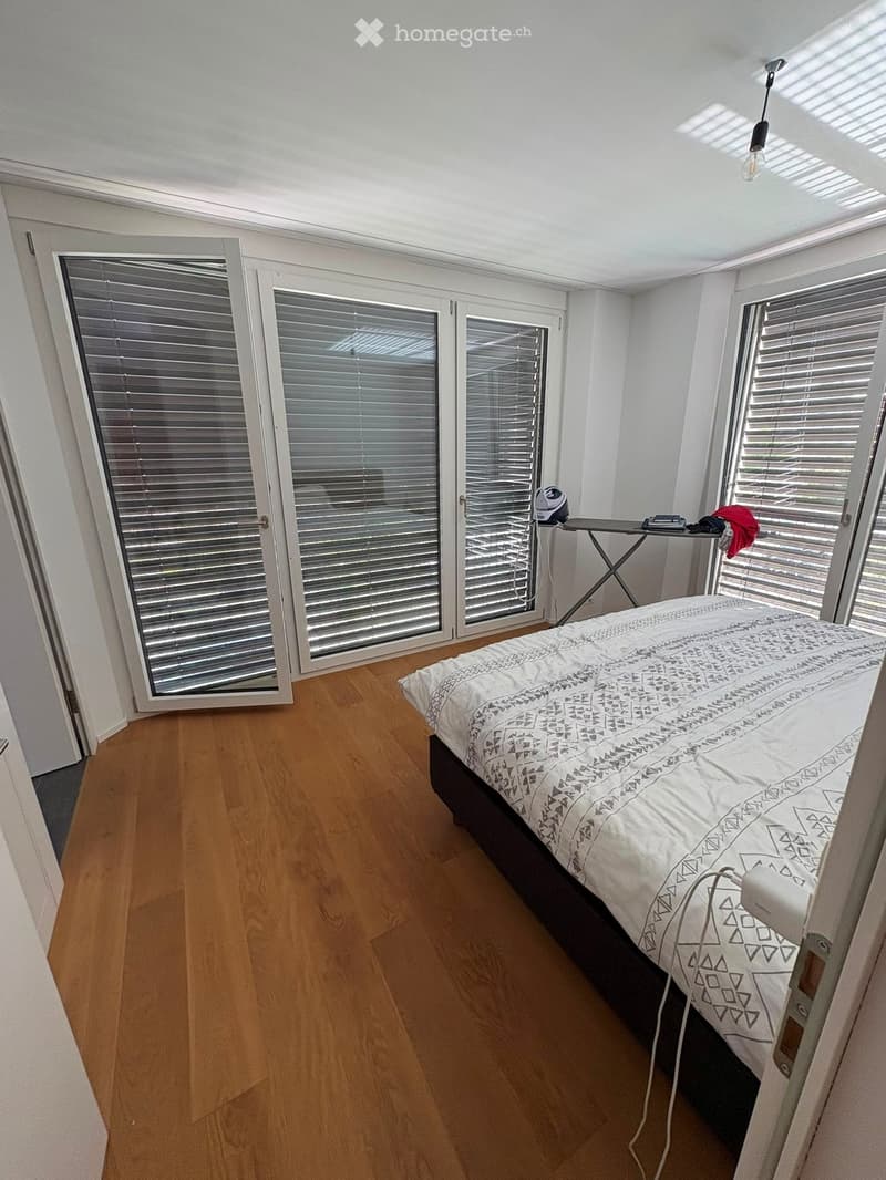 Bright bedroom with wooden flooring, large windows with blinds, and a bed with patterned bedding. An ironing board and iron are placed near the window.