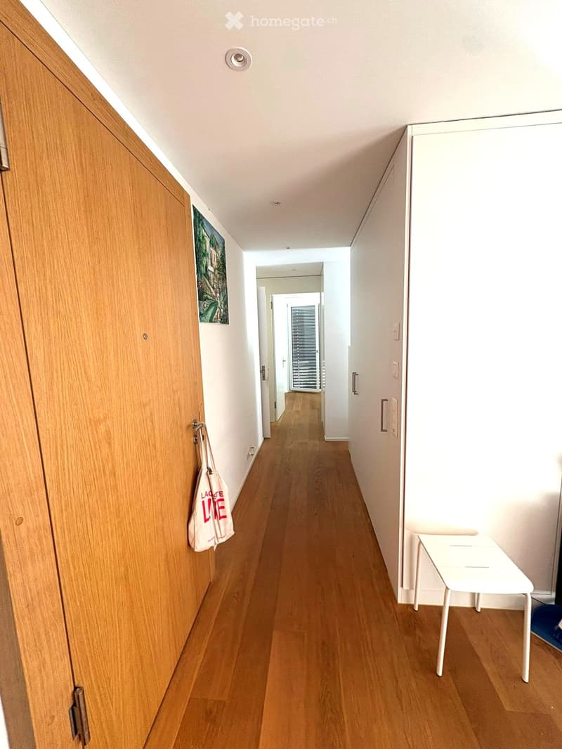 A narrow hallway with wooden flooring, white walls, and a small white table on the right side. A wooden door is on the left, and a painting hangs on the wall above it. A white bag is hanging on the door handle.