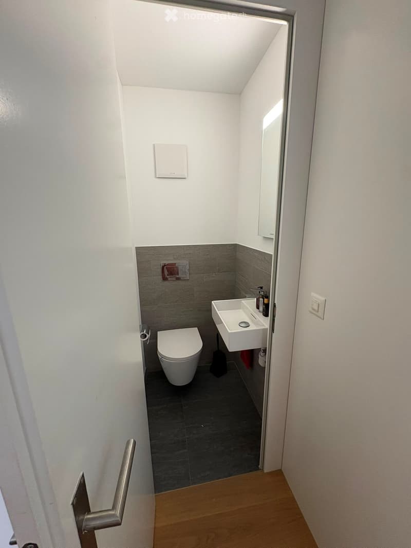 Modern, compact bathroom with white walls, grey tiled lower half, and dark grey floor. Features a toilet, sink with a mirror above, and a light switch on the wall.